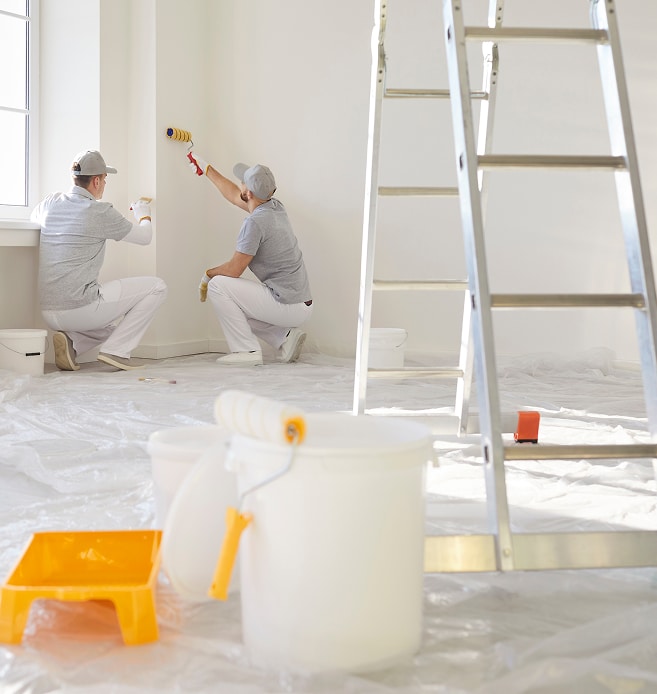 Deux hommes peintres professionnels rénovent un intérieur, peignant des murs blancs avec rouleaux. Équipement de peinture au sol.