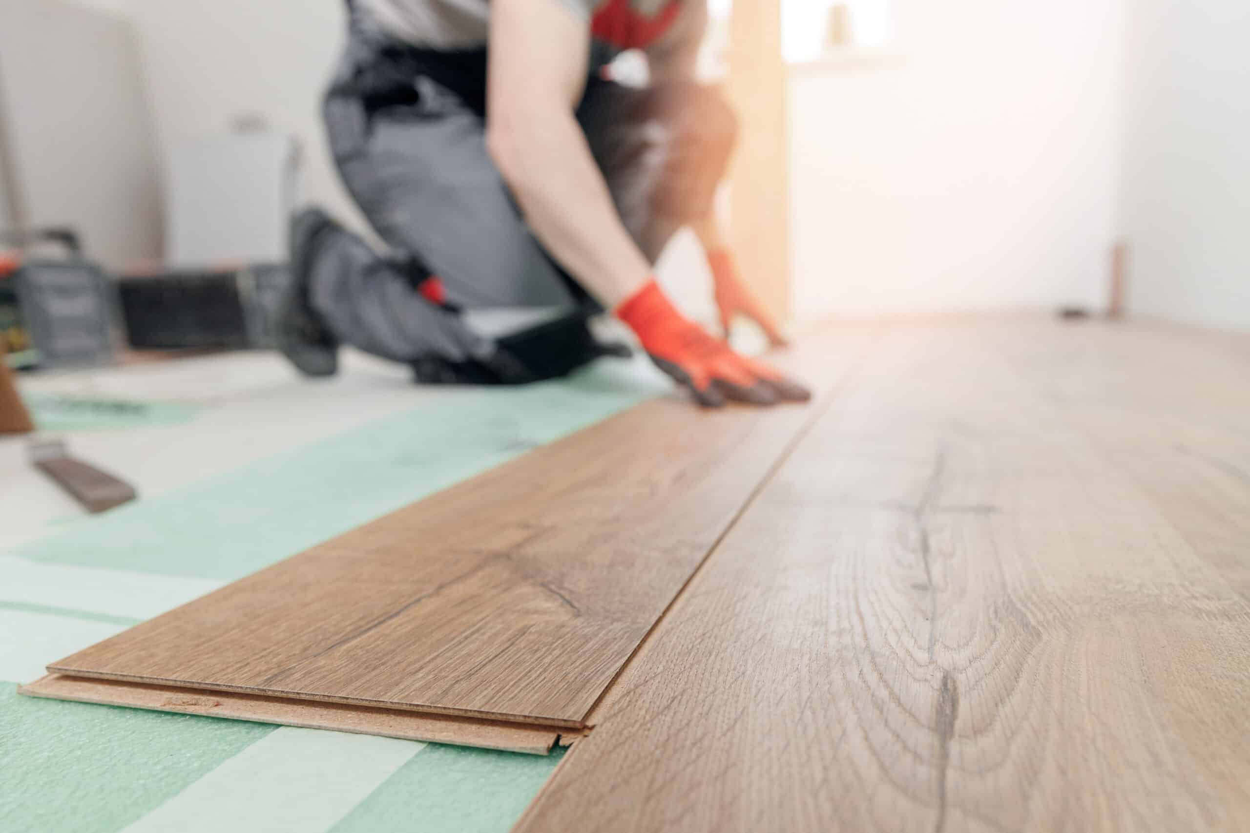 Un poseur installe du parquet stratifié brun sur une sous-couche verte. Il est accroupi et porte des gants. Rénovation de sol.